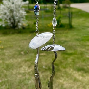 Può includere: Due ornamenti pendenti realizzati con cucchiai in metallo argentato a forma di fungo. Ogni pezzo presenta la coppa del cucchiaio come cappello e il manico come gambo, sospesi da una catenella d'argento con un dettaglio in perla di vetro.