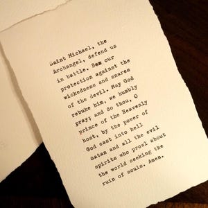May include: A rectangular piece of off-white, textured paper with deckled edges featuring black typewriter-style text. The printed prayer reads: Saint Michael, the Archangel, defend us in battle. Be our protection against the wickedness and snares of the devil. May God rebuke him, we humbly pray; and do thou, O prince of the Heavenly host, by the power of God cast into hell Satan and all the evil spirits who prowl about the world seeking the ruin of souls. Amen.
