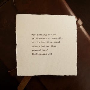 May include: A square piece of off-white paper with deckled edges featuring black typewriter-style text that reads: "Do nothing out of selfishness or conceit, but in humility count others better than yourselves." Philippians 2:3.