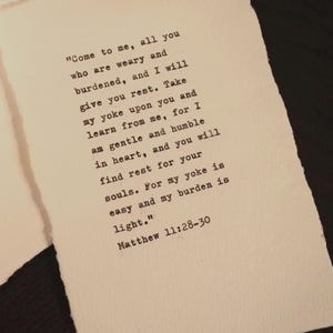 May include: A rectangular piece of off-white paper with rough, deckled edges featuring black typewriter-style text. The text reads: "Come to me, all you who are weary and burdened, and I will give you rest. Take my yoke upon you and learn from me, for I am gentle and humble in heart, and you will find rest for your souls. For my yoke is easy and my burden is light." Matthew 11:28-30.