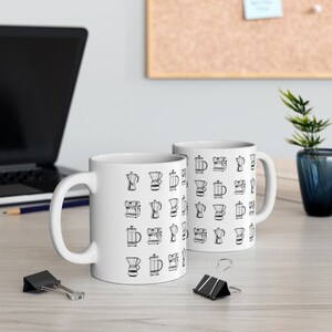 Op de afbeelding: Twee witte keramische mokken met zwarte contouren van verschillende koffiezetmethoden, zoals een French Press, een pour over en een espressomachine, staan op een houten tafel.