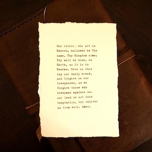 May include: A rectangular piece of off-white paper with deckled edges featuring black typewriter-style text of the Lord's Prayer. The paper is placed on top of a dark brown leather-bound book with a visible strap closure.