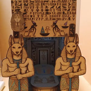 Pode incluir: Diorama egípcio de madeira detalhado com duas figuras de Anúbis segurando bastões, ladeando um sarcófago. O fundo inclui hieróglifos, uma cabeça de faraó e outros símbolos egípcios. O diorama é pintado em tons de castanho, bege e azul.