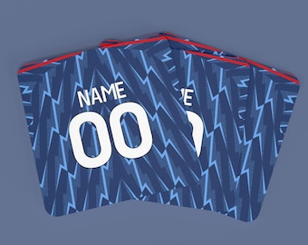 May include: A stack of blue and light blue jersey-style coasters with a lightning bolt pattern. The coasters have the text "NAME" and the number "00" in white. The edges are trimmed with red.