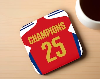 Può includere: Sottobicchiere quadrato rosso con design a maglia. Il sottobicchiere presenta la scritta "CHAMPIONS" in lettere dorate, insieme al numero "25". Accenti bianchi e blu incorniciano il design. Una tazza bianca con liquido scuro è vicina.