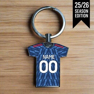 Op de afbeelding: Een zilveren sleutelhanger met een voetbalshirtontwerp. Het blauwe shirt heeft witte bliksemflitspatronen en het nummer "00". Boven het nummer staat het woord "NAME". De tekst "25/26 SEASON EDITION" staat op een zwarte banner.