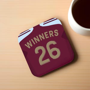 Peut inclure: Un dessous de verre marron avec le mot "WINNERS" et le chiffre "26" en or. Le dessous de verre a un motif bleu clair et blanc en haut. Une tasse blanche remplie d'un liquide foncé est en arrière-plan.