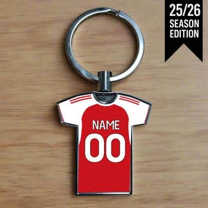 May include: A red and white jersey-shaped keychain with the text "NAME" and the number "00" on the front. The keychain has a silver ring and the text "25/26 SEASON EDITION" on a black banner.