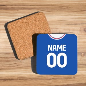 May include: A blue square coaster with the word "NAME" and the number "00" in white. The coaster has a red and white trim at the top, resembling a jersey. A separate cork coaster is next to it.
