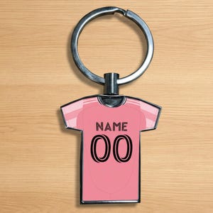 Op de afbeelding: Roze sleutelhanger in de vorm van een shirt met een zilveren sleutelring. Het shirt heeft het woord "NAME" boven het nummer "00" in zwart. De sleutelhanger ligt op een houten oppervlak.