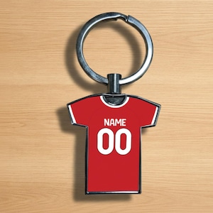 Könnte beinhalten: Ein roter schlüsselanhänger in Trikotform mit dem Text "NAME" und der Nummer "00" in Weiß. Der Schlüsselanhänger hat einen silbernen Metallring und einen schwarzen Verbinder.