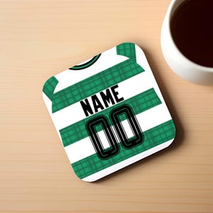 Peut inclure: Un dessous de verre carré avec un motif à carreaux vert et blanc rappelant un maillot de sport. Le motif du maillot comprend le mot "NAME" et le numéro "00" en noir. Une tasse à café blanche remplie d'un liquide foncé est à proximité sur une surface en bois.