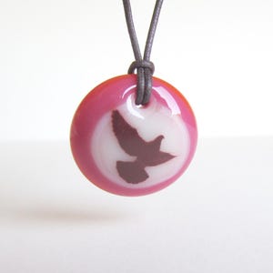 K&ouml;nnte beinhalten: Eine runde Anh&auml;nger Halskette mit rosa und wei&szlig;em Design. Der Anh&auml;nger zeigt eine dunkelbraune Vogelsilhouette vor wei&szlig;em Hintergrund, umgeben von einem rosa Rand. Die Halskette hat eine graue Kordel.