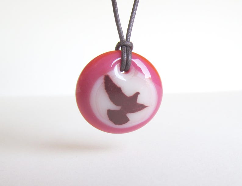 K&ouml;nnte beinhalten: Eine runde Anh&auml;nger Halskette mit rosa und wei&szlig;em Design. Der Anh&auml;nger zeigt eine dunkelbraune Vogelsilhouette vor wei&szlig;em Hintergrund, umgeben von einem rosa Rand. Die Halskette hat eine graue Kordel.