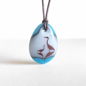 May include: A teardrop-shaped glass pendant featuring a white center with a brown silhouette of two birds and their reflection, bordered by a bright blue edge. The pendant hangs from a dark brown cord threaded through a hole at the top.