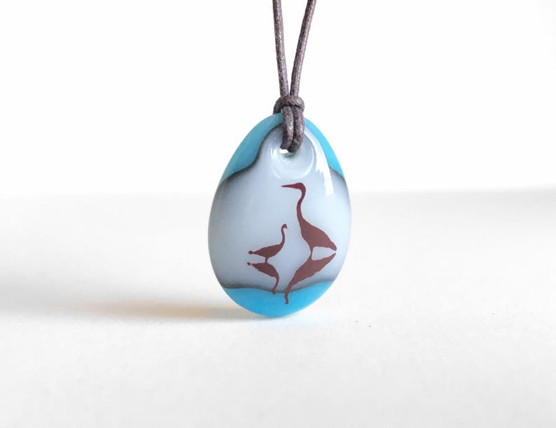 May include: A teardrop-shaped glass pendant featuring a white center with a brown silhouette of two birds and their reflection, bordered by a bright blue edge. The pendant hangs from a dark brown cord threaded through a hole at the top.