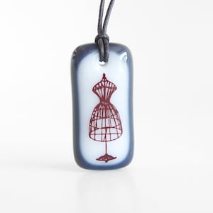 May include: A gray glass pendant necklace with a white background and a red outline of a dress form. The necklace is on a gray cord.