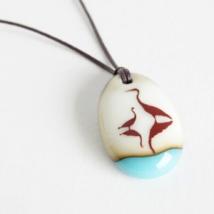 May include: An oval pendant on a dark brown cord featuring a smooth off-white surface with a dark red silhouette of two birds. The bottom section of the pendant is a solid, bright turquoise blue color, creating a distinct contrast with the upper area.