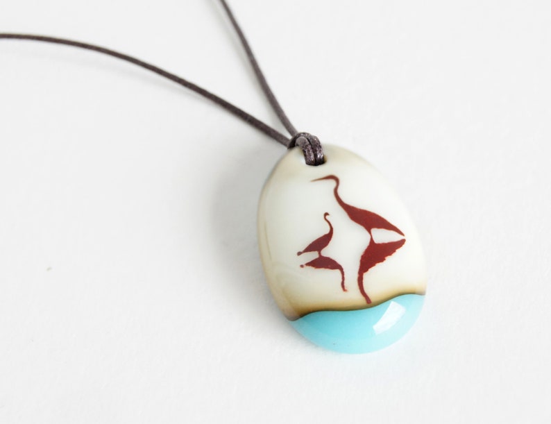 May include: An oval pendant on a dark brown cord featuring a smooth off-white surface with a dark red silhouette of two birds. The bottom section of the pendant is a solid, bright turquoise blue color, creating a distinct contrast with the upper area.