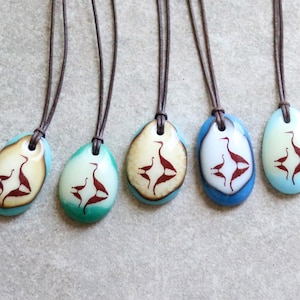 May include: Five oval-shaped pendants on dark brown leather cords. Each pendant features a smooth, polished surface in shades of blue, teal, or cream, decorated with a dark brown silhouette graphic of two birds facing each other in a mirrored design.