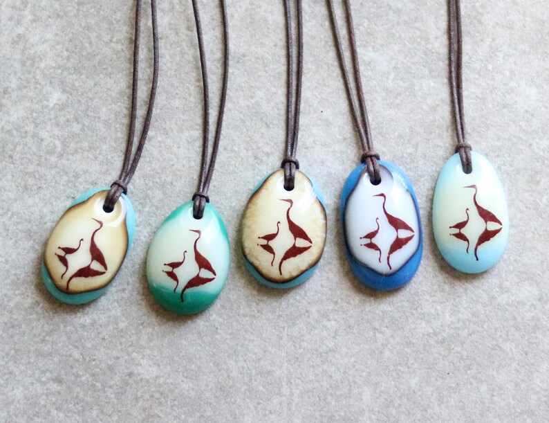 May include: Five oval-shaped pendants on dark brown leather cords. Each pendant features a smooth, polished surface in shades of blue, teal, or cream, decorated with a dark brown silhouette graphic of two birds facing each other in a mirrored design.