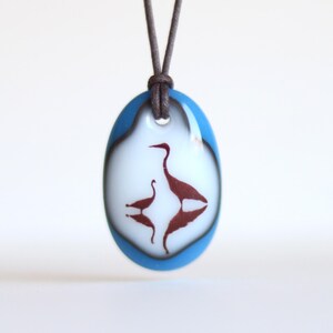 May include: An oval glass pendant featuring a white center with a dark red silhouette of two birds, one larger and one smaller, with their reflections below. The pendant has a bright blue border and hangs from a dark brown cord necklace.