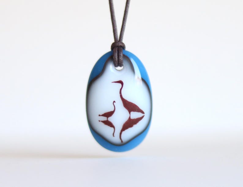 May include: An oval glass pendant featuring a white center with a dark red silhouette of two birds, one larger and one smaller, with their reflections below. The pendant has a bright blue border and hangs from a dark brown cord necklace.