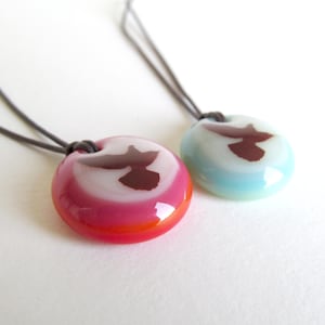 K&ouml;nnte beinhalten: Zwei runde Anh&auml;nger-Halsketten. Einer ist rosa und wei&szlig;, der andere hellblau und wei&szlig;. Jeder hat ein dunkelbraunes abstraktes Design und eine dunkelgraue Kordel. Die Anh&auml;nger sind gl&auml;nzend und scheinen aus Glas oder Harz zu bestehen.