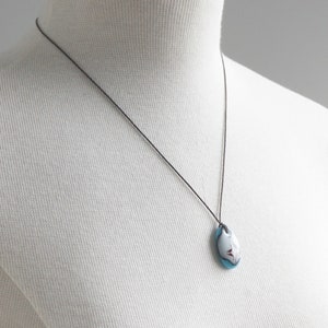 May include: A necklace featuring an oval-shaped pendant with a smooth, glossy finish. The pendant displays a color palette of white, light blue, and dark red accents. It is suspended from a thin, dark brown cord that rests against the white mannequin.