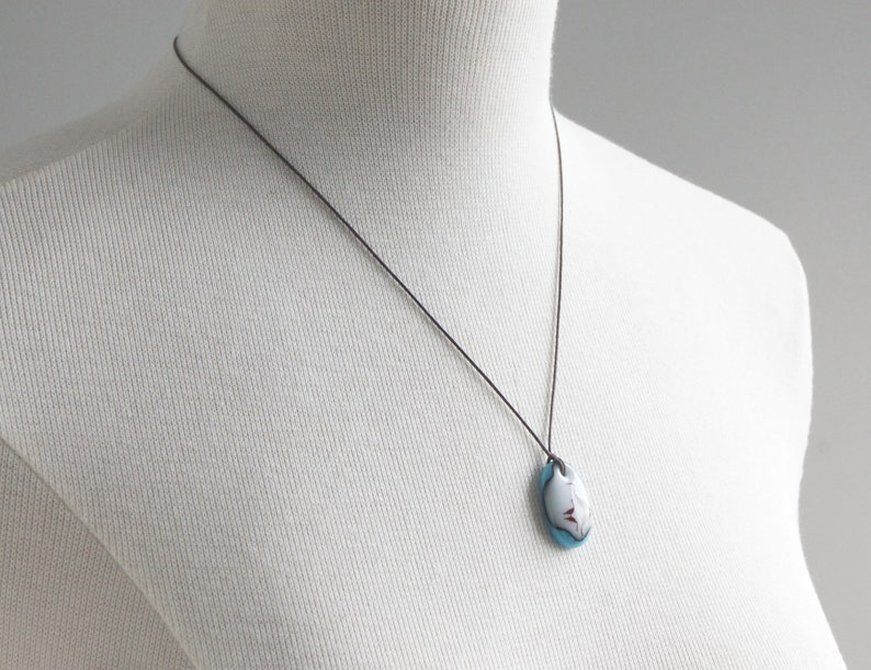 May include: A necklace featuring an oval-shaped pendant with a smooth, glossy finish. The pendant displays a color palette of white, light blue, and dark red accents. It is suspended from a thin, dark brown cord that rests against the white mannequin.