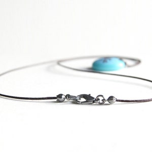 May include: A necklace featuring a thin dark brown cord and a metallic lobster claw clasp. The clasp is flanked by two small metallic beads. A smooth, rounded, bright turquoise blue pendant is attached to the cord, resting out of focus.