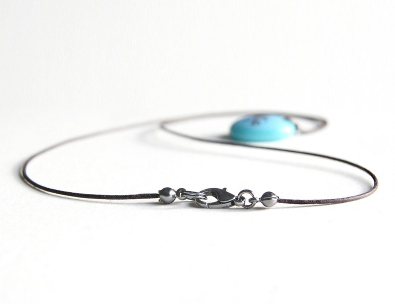 May include: A necklace featuring a thin dark brown cord and a metallic lobster claw clasp. The clasp is flanked by two small metallic beads. A smooth, rounded, bright turquoise blue pendant is attached to the cord, resting out of focus.