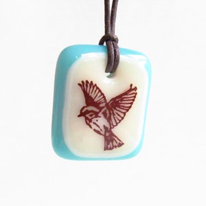K&ouml;nnte beinhalten: Eine quadratische Anh&auml;nger-Halskette mit braunem Lederband. Der Anh&auml;nger hat einen hellblauen Rand und eine cremefarbene Mitte mit einer braunen Illustration eines Vogels im Flug. Die Halskette ist ein Accessoire zum Thema Natur.