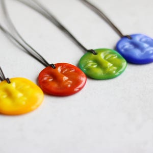 Könnte beinhalten: Vier Halsketten mit farbenfrohen Anhängern. Jeder Anhänger ist ein runder, glänzender Anhänger mit einem geformten Gesicht. Die Anhänger sind gelb, rot-orange, grün und blau. Die Halsketten haben dünne, dunkle Kordeln.