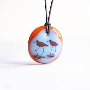 Könnte beinhalten: Ein runder orangefarbener Glasanhänger mit blauem Hintergrund, auf dem zwei braune Silhouetten von Vögeln im Wasser stehen. Der Anhänger ist an einer schwarzen Schnur befestigt.