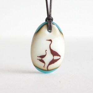 May include: An oval glass pendant featuring a cream-colored center with a dark brown silhouette of two birds facing each other. The pendant is framed by a translucent light blue border and hangs from a dark brown leather cord through a top hole.
