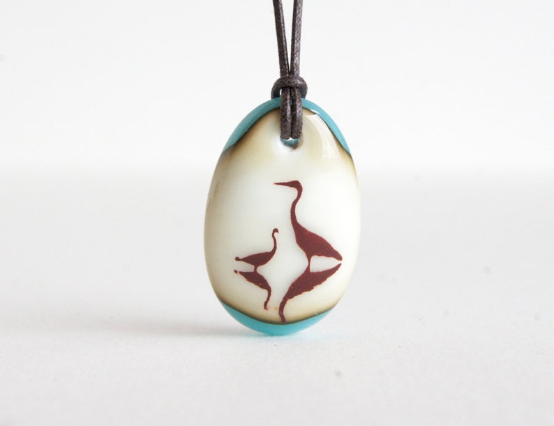 May include: An oval glass pendant featuring a cream-colored center with a dark brown silhouette of two birds facing each other. The pendant is framed by a translucent light blue border and hangs from a dark brown leather cord through a top hole.