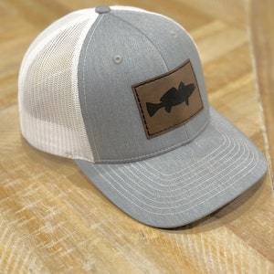May include: A gray and white mesh trucker hat with a brown leather patch featuring a black silhouette of a fish.