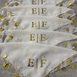 Puede incluir: Pañuelos blancos con ribete dorado y adornos de monedas doradas. Cada pañuelo tiene las letras doradas "E|F" impresas. Los pañuelos están dispuestos en fila sobre una superficie gris.