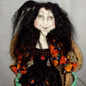 May include: A handmade doll with long black hair, wearing a black dress with orange and green pumpkin accents. The doll is sitting in a wicker chair.