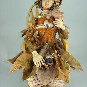 Może przedstawiać: Ręcznie robiona lalka z brązowymi kręconymi włosami, ubrana w brązową i złotą sukienkę, trzymająca kota i włóczkę w koszyku. Lalka siedzi na drewnianym krześle.