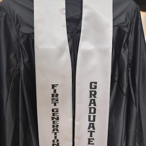 May include: A black graduation gown with a white sash. The sash features the words "FIRST GENERATION" and "GRADUATE" in black block lettering. The gown is suspended from a hanger.