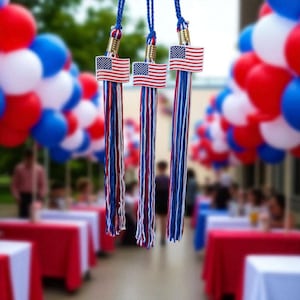 May include: Three graduation tassels with American flags. Each tassel has red, white, and blue cords, with a small flag emblem at the top. The background includes red, white, and blue balloons and tables.
