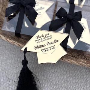 Op de afbeelding: Afstudeerbedankjes met een goudkleurig baretontwerp en een zwarte kwast. Elk bedankje is verpakt in een doorzichtige doos en vastgebonden met een zwart satijnen lint. De tekst op de baret luidt "Bedankt voor het vieren van mijn afstuderen" en bevat een naam en "Klasse van 2024."