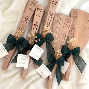 Puede incluir: Un conjunto de abanicos de mano de madera con intrincados diseños calados. Cada abanico está adornado con un lazo de cinta verde oscuro, flores secas y una pequeña etiqueta. Los abanicos están dispuestos sobre un fondo de tela blanca, creando una exhibición elegante.