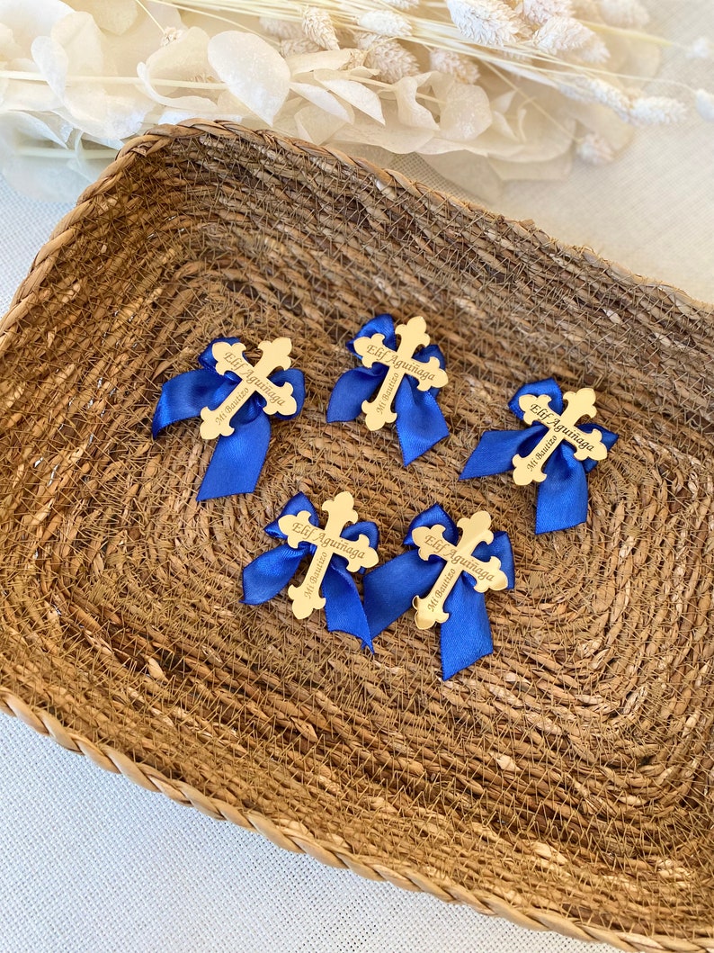 K&ouml;nnte beinhalten: Sechs goldfarbene Kreuze mit blauen Schleifen. Jedes Kreuz tr&auml;gt den Namen "Elif Aguinaga" und "Mi Bautizo" eingraviert.
