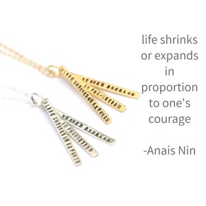 May include: Two necklaces with pendants that have the quote "Life shrinks or expands in proportion to one's courage" by Anais Nin engraved on them. The necklaces are made of gold and silver.