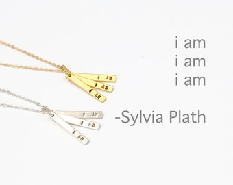May include: Two necklaces with three small gold and silver bars hanging from them. Each bar has the words "I AM" engraved on it. The text "i am i am i am - Sylvia Plath" is written below the necklaces.