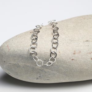 May include: A silver chain bracelet with a series of interconnected, open circular links. The bracelet is displayed on a light gray stone, creating a contrast between the shiny metal and the natural texture of the stone. The bracelet is a piece of jewelry.