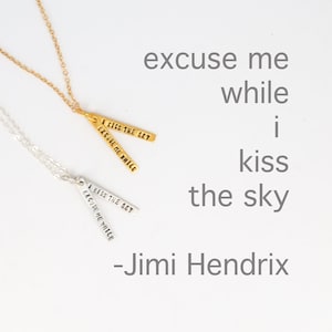 Op de afbeelding: Twee zilveren en gouden kettingen met een hanger waarop aan beide kanten "I kiss the sky, excuse me while" staat. De kettingen liggen op een witte achtergrond met de tekst "excuse me while I kiss the sky - Jimi Hendrix".